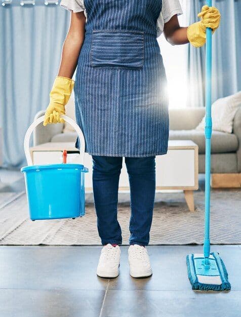 Residential cleaner in apron and yellow gloves with mop and bucket in a bright modern living room